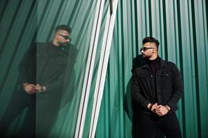 El hombre árabe de barba rica en moda viste una chaqueta negra de jeans y gafas de sol posadas contra una pared verde con ventanas. chico modelo árabe elegante, exitoso y de moda. foto