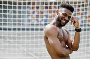 guapo sexy afroamericano musculoso torso desnudo hombre en overoles de jeans posó en la hierba verde contra las puertas de fútbol del campo del estadio. retrato de hombre negro de moda. foto