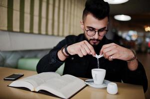 Arab man wear on black jeans jacket and eyeglasses sitting in cafe, read book and drink coffee. Stylish and fashionable arabian model guy. Pour sugar at cup. photo