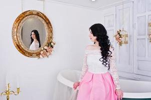 Young brunette girl in pink skirt and white blouse posed indoor against room with decor bath and mirror. photo