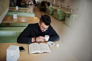 Arab man wear on black jeans jacket and eyeglasses sitting in cafe, read book and drink coffee. Stylish and fashionable arabian model guy. photo