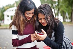 Portrait of two young beautiful indian or south asian teenage girls in dress sitting on bench and use mobile phone. photo