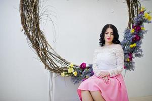 jovencita morena con falda rosa y blusa blanca posada en el interior contra una gran corona decorada. foto