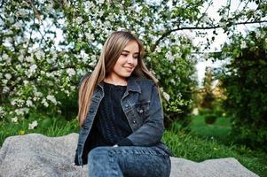 jovencita morena en jeans sentada en tela escocesa contra el árbol de flores de primavera. foto