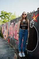 elegante chica hipster casual con gorra, gafas de sol y jeans desgastados contra una gran pared de graffiti con una gran bomba tnt. foto