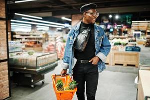 Elegante hombre afroamericano casual con chaqueta de jeans y boina negra sosteniendo una canasta con piñas en la sección orgánica de frutas del supermercado. foto