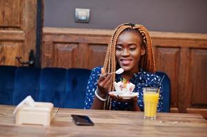 Portrait of beautiful young african business woman with dreadlocks, wear on blue blouse and skirt, sitting in cafe with ice cream and pineapple juice. photo