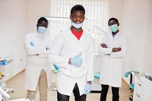 Three african american male doctor with crossed arms in dental clinic. Show thumb up. photo