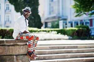 Stylish african american man in white shirt and colored pants with hat and glasses posed outdoor. Black fashionable model boy. photo