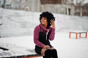 mujer afroamericana de pelo rizado posó en el día de invierno, sentada en un banco. foto