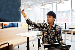 joven autónomo indio sentado con una computadora portátil abierta en un café de comida rápida, un apuesto hombre asiático sostiene el plato con el pedido y espera al camarero en una cómoda cafetería. foto