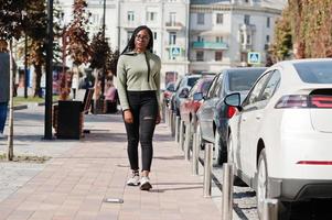 City portrait of positive young dark skinned female wearing green hoody and eyeglasses walking at car parking. photo