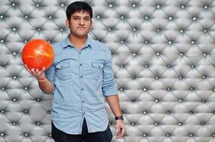 Stylish asian man in jeans shirt standing with bowling ball at hand against silver wall background. photo