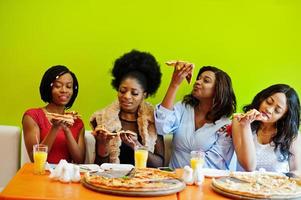 Four young african girls in bright colored restaurant eating pizza slices in hands. photo