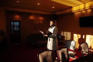 Handsome african american man posing  inside room with sunlight shadows in black hat and beige coat. photo