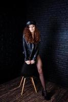 chica morena de pelo rizado y piernas largas con chaqueta de cuero negro con traje de baño y gorra posada en el estudio en una silla contra una pared de ladrillo oscuro. foto