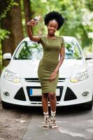 African american woman posed against white car in forest road and looking at mobile phone. photo