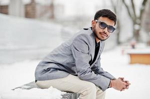 un joven indio informal con chaqueta plateada y gafas de sol posó en el día de invierno y se sentó en un banco. foto