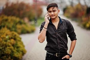 hombre indio con camisa marrón posó al aire libre y hablando por teléfono móvil. foto