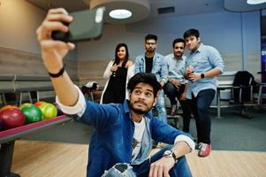Group of five south asian peoples having rest and fun at bowling club. Making selfie by phone and holding cold soda drinks from glass bottles. photo