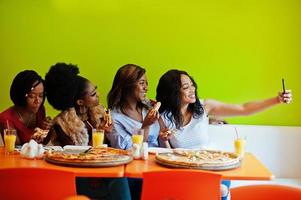 Four young african girls in bright colored restaurant eating pizza, having fun together and making selfie on phone. photo