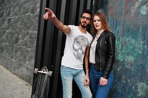 Cool pareja multirracial posando contra la pared con longboard. El novio árabe le muestra algo a su chica europea. foto