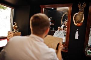 Young bearded man getting haircut by hairdresser while sitting in chair at barbershop. Barber soul. photo