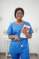 retrato de una joven pediatra afroamericana feliz con uniforme azul y estetoscopio con libros en las manos. salud, medicina, especialista en medicina - concepto. foto