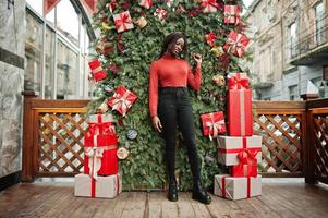 Portrait of a curly haired african woman wearing fashionable red turtleneck posing against christmas decorations, new year eve theme. photo