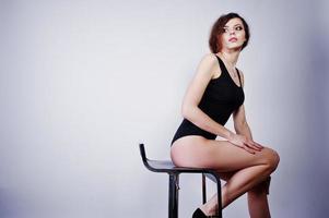 Brunette curly haired long legs girl in black swimsuit posed on chair at studio against white background. photo