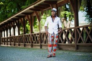 Stylish african american man in white shirt and colored pants with hat and glasses posed outdoor. Black fashionable model boy. photo