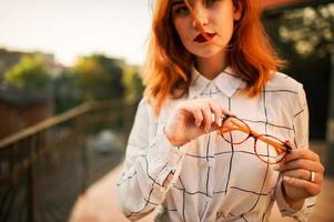 Close up photo of eyeglasses holding redhaired  woman, wear white blouse.