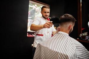 Young bearded man getting haircut by hairdresser while sitting in chair at barbershop. Barber soul. photo