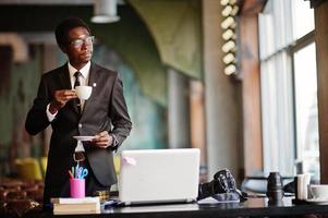 African american photographer paparazzi man wear on black suit and glasses at office with laptop, drinking morning coffee before work day. photo