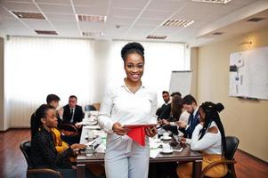 Face of handsome african american business woman, holding tablet on the background of business peoples multiracial team meeting, sitting in office table. photo