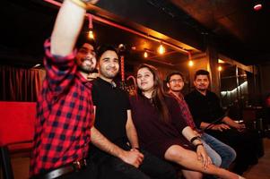 grupo de amigos indios sentados en el lounge bar, divirtiéndose y descansando, fumando narguile y mirando teléfonos móviles para hacer selfie. foto