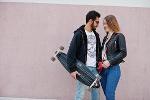 Cool pareja multirracial posando contra la pared de color rosa con longboard. foto