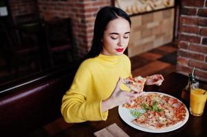 chica morena divertida en suéter amarillo comiendo pizza en el restaurante. foto