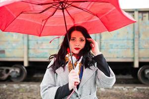 chica morena con abrigo gris con paraguas rojo en la estación de tren. foto