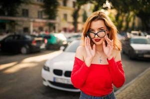 Attractive redhaired woman in eyeglasses, wear on red blouse and jeans skirt posing at street against white sport car. photo