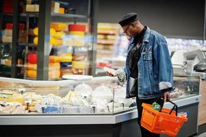 Stylish casual african american man at jeans jacket and black beret holding basket, standing near cheese fridge and shopping at supermarket. photo