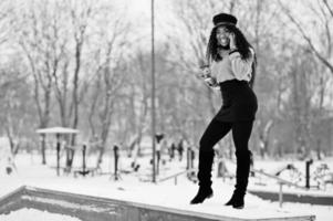 African american woman in black skirt, brown sweater and cap posed at winter day against snowy background, holding cup of coffee and speaking on phone. photo