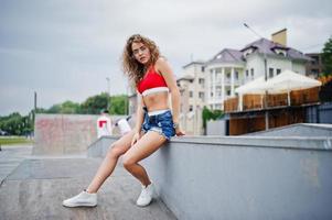 Sexy curly model girl in red top, jeans denim shorts, eyeglasses and sneakers posed at skatepark. photo