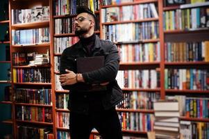 estudiante árabe alto e inteligente, vestido con chaqueta negra de jeans y anteojos, en la biblioteca con un libro en las manos. foto