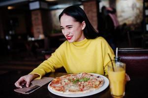 Funny brunette girl in yellow sweater eating pizza at restaurant. photo