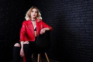 retrato de estudio de una chica rubia con chaqueta de cuero roja posada en una silla contra una pared de ladrillo. foto