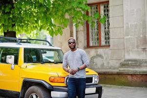 Stylish african american boy on gray sweater and black sunglasses posed on street against yellow car. Fashionable black business man. photo