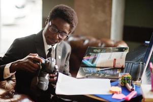 African american photographer paparazzi man wear on black suit and glasses sitting at office with camera and working behind laptop. photo