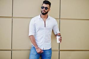 Elegante modelo de hombre árabe alto con camisa blanca, jeans y gafas de sol en la calle de la ciudad. barba chico árabe atractivo con una taza de café contra la pared dorada. foto