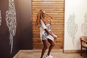 hermosa mujer africana con ropa informal elegante posando contra la pared de alas de ángel. ella moviendo sus rastas de pelo. foto